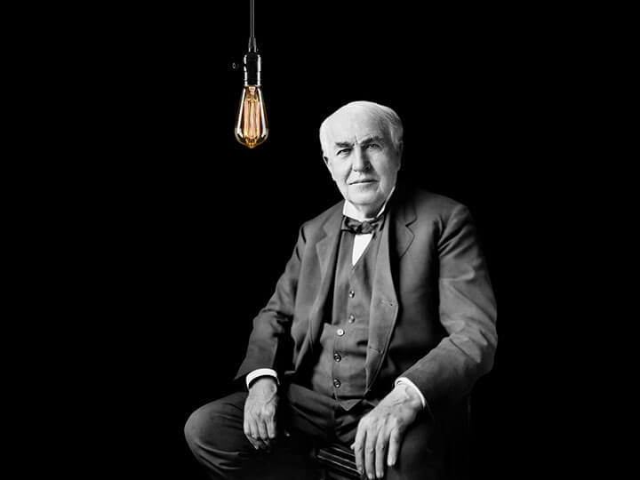 Thomas Edison sitting next to an Edison lightbulb with a black background