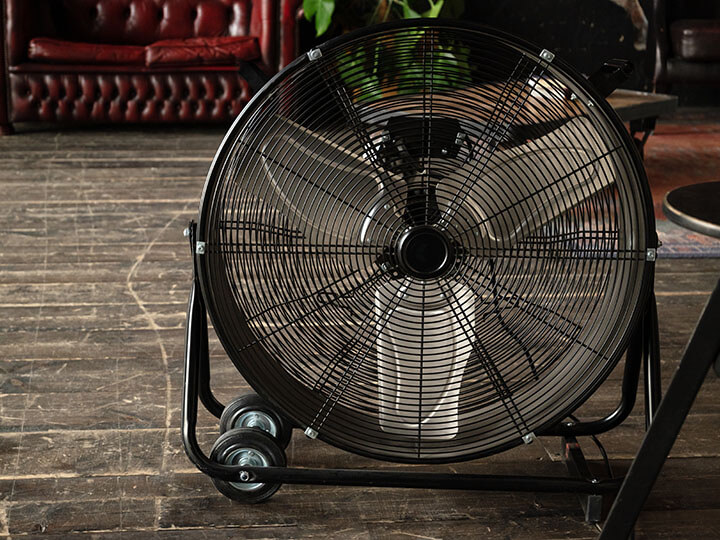 Round black cooling fan with wheels on a rustic wooden floor in room with a red armchair in the background